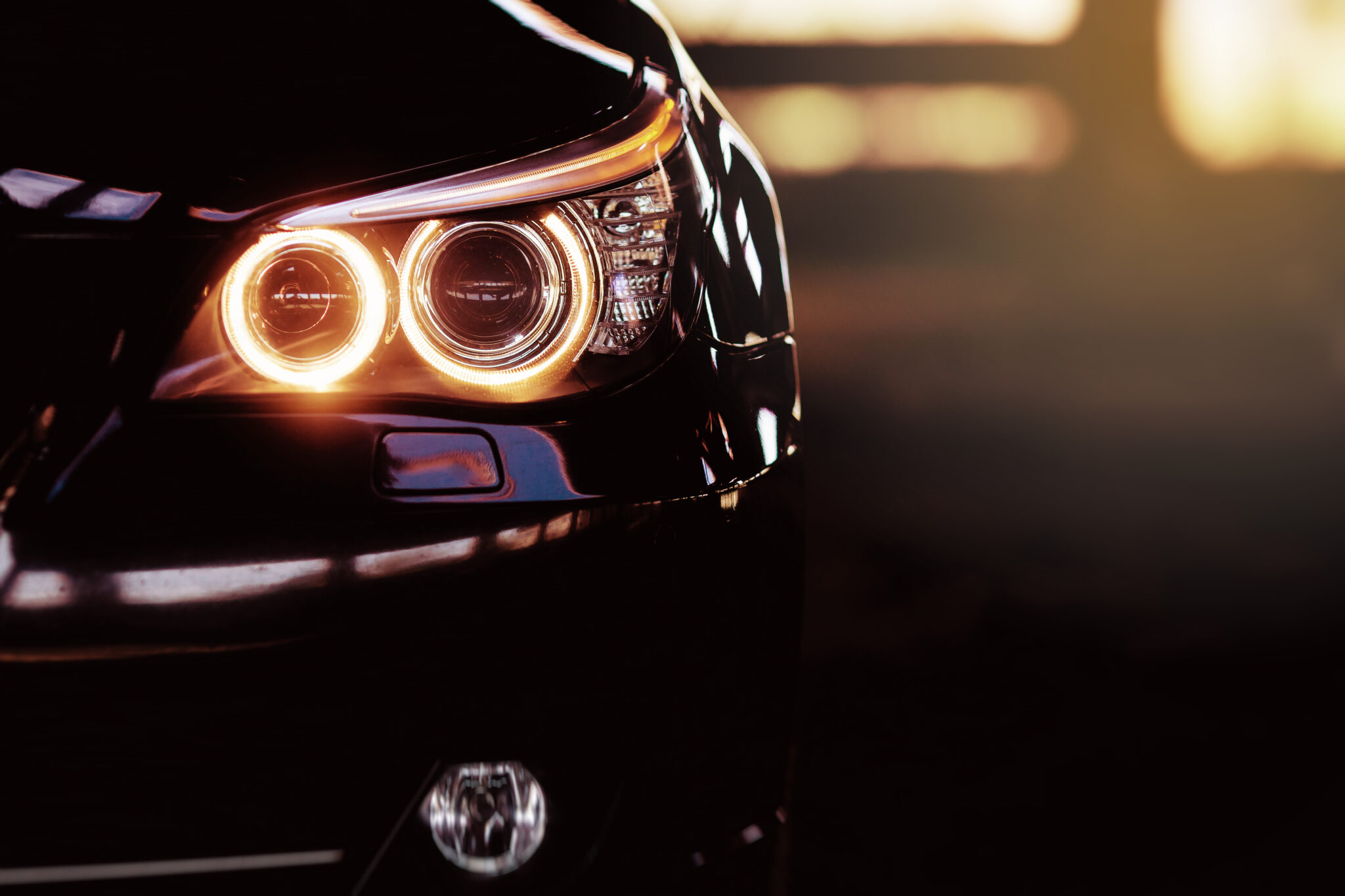 Close-up of a car’s front headlight illuminated at night, with warm light reflecting off the glossy black vehicle.