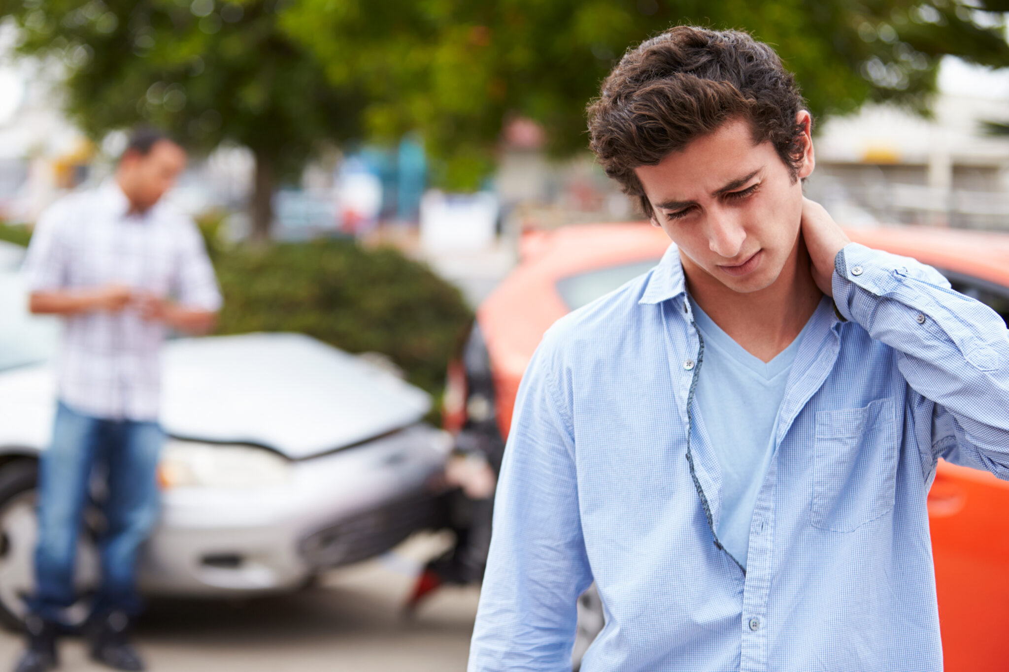 Young man holding his neck in pain at the scene of a car accident with damaged vehicles in the background