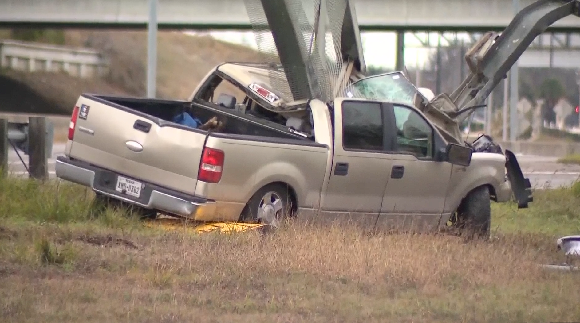 Severely damaged pickup truck off the roadway after a crash, with the truck bed crushed and debris visible near an overpass.