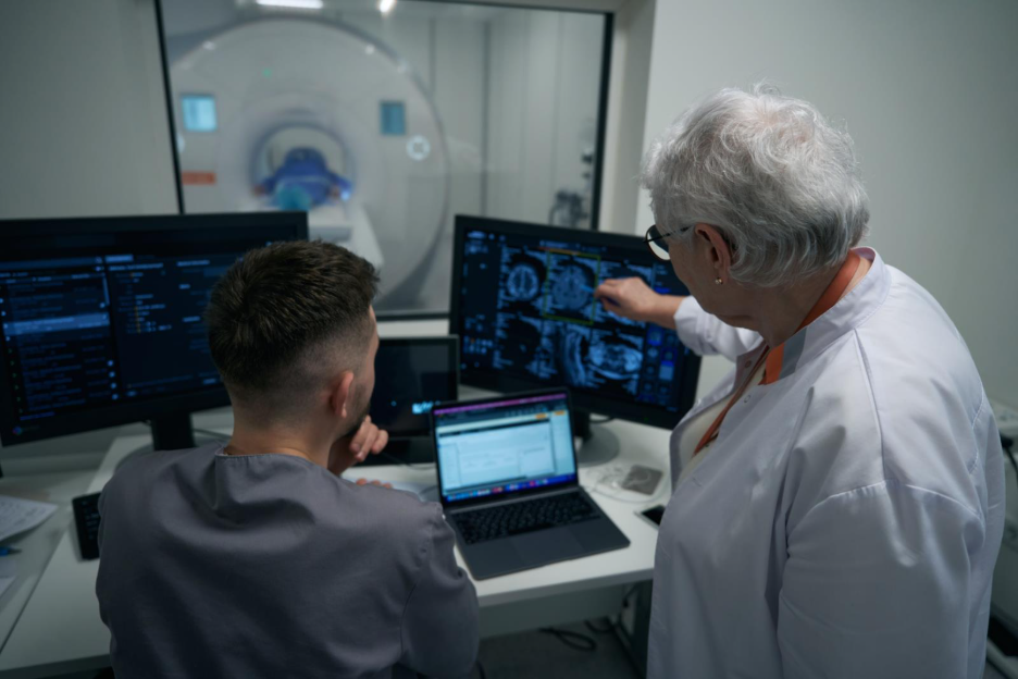 Two medical professionals reviewing brain imaging scans on computer monitors in a radiology control room.