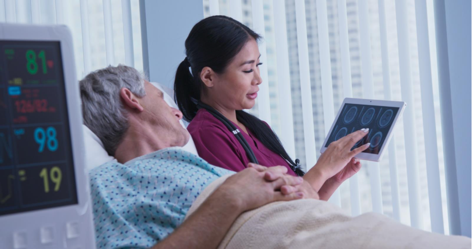 Nurse showing medical scan results on a tablet to an older patient lying in a hospital bed.