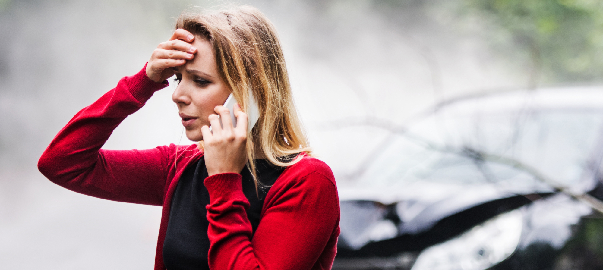 Woman talking on cell phone, holding her head in shock and frustration after a car crash.