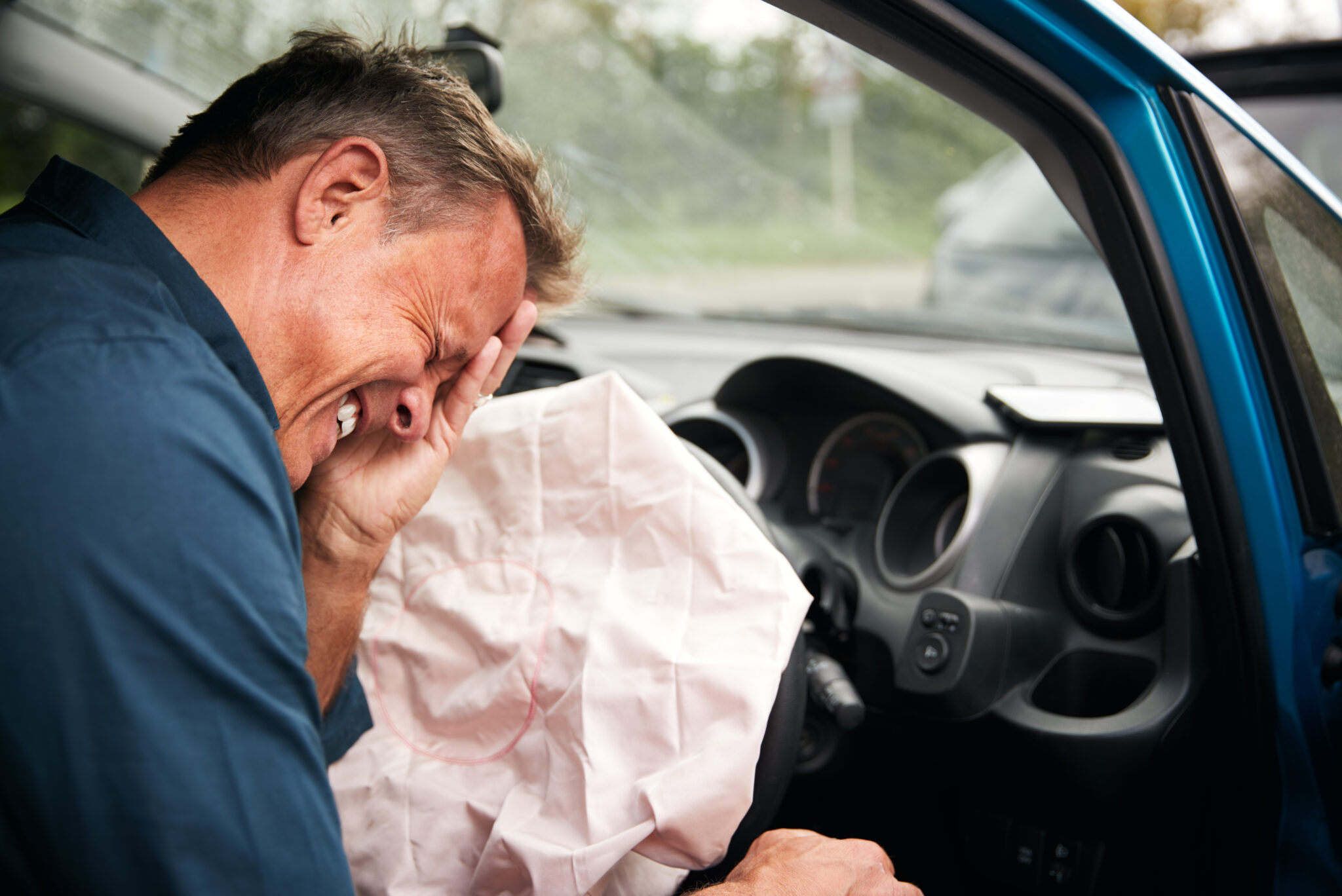 Injured driver holding his face beside a deployed airbag inside a blue car after a collision.