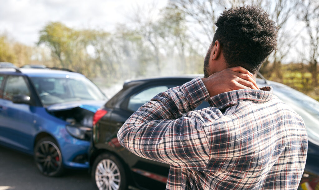Man standing outside two crashed cars on a road, holding his neck in pain, showing possible whiplash injury.