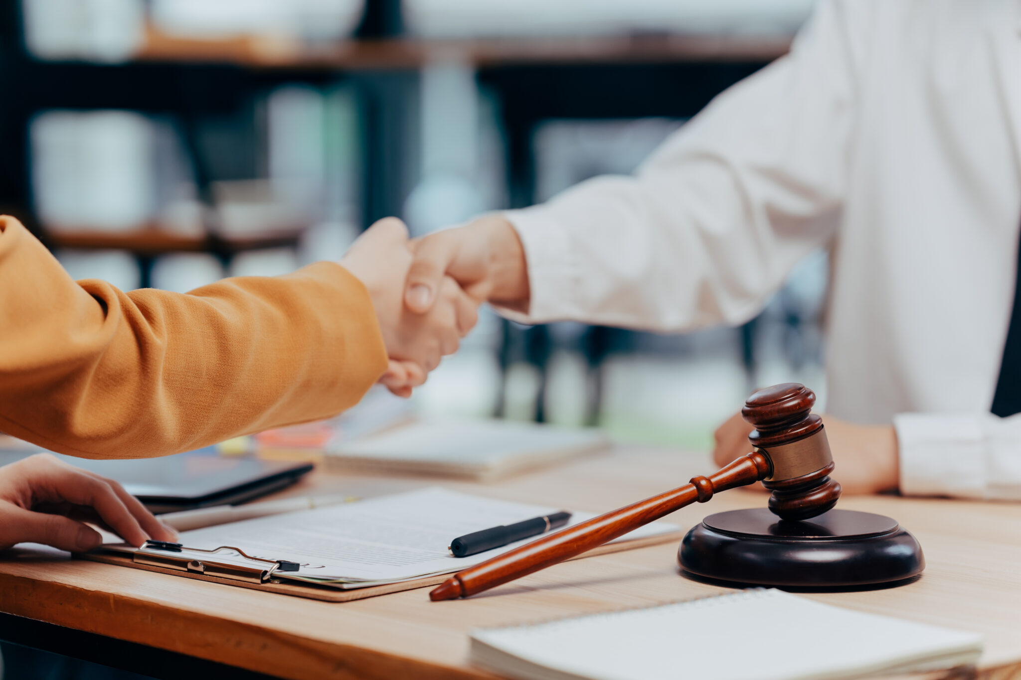 Two people shake hands across a desk with a judge’s gavel, clipboard, and documents visible, symbolizing a legal agreement or settlement.