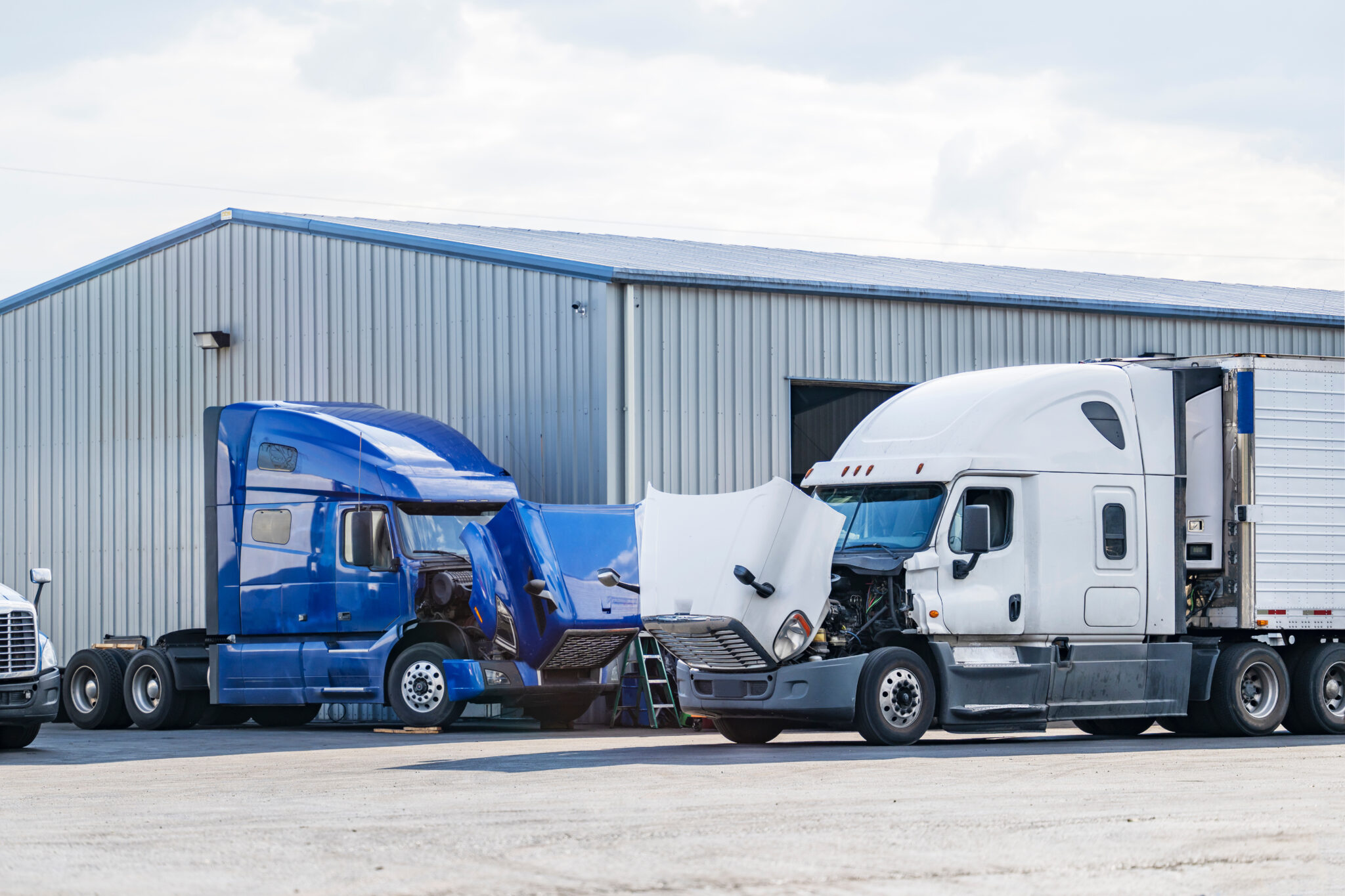 Bonnet industrial freight broken big rig semi trucks with open hoods standing on the parking lot of the repair shop for engine maintenance and inspection and necessary current repairs