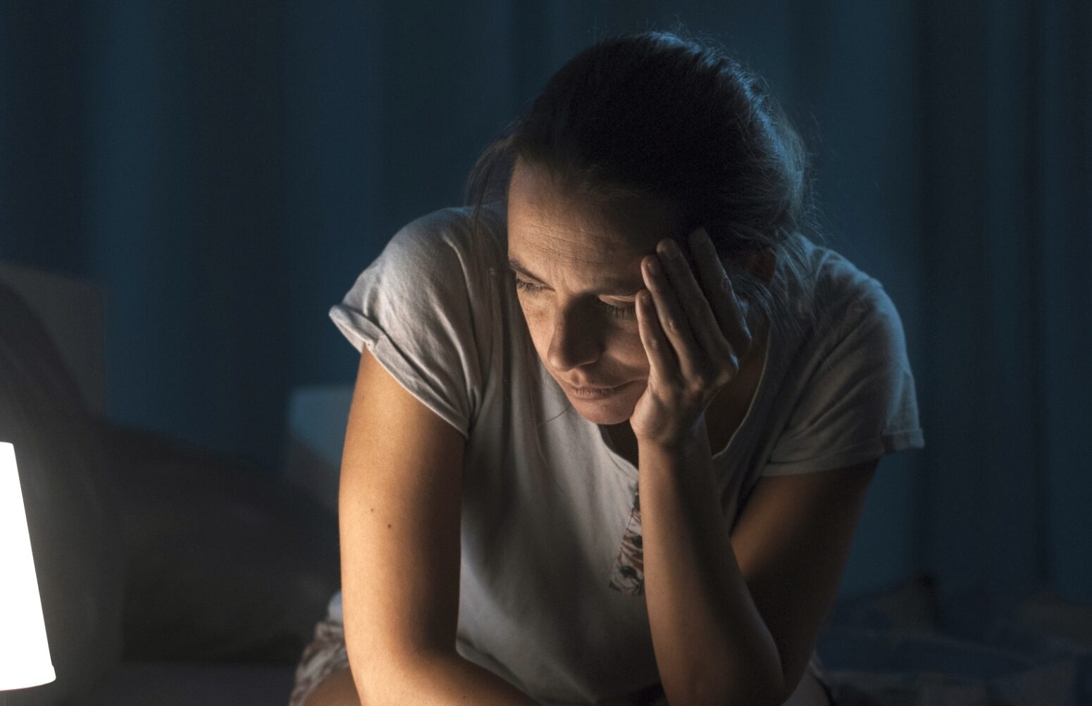 A woman sitting in a darkened room with her head in her hand.