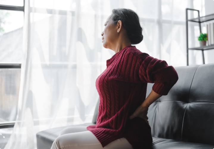 An older asian woman sitting on her couch holding her back in pain