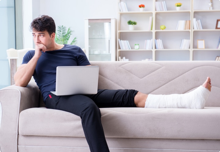 White male in deep thoughts with a cast on his left leg while sitting on a coach with his laptop on his lap