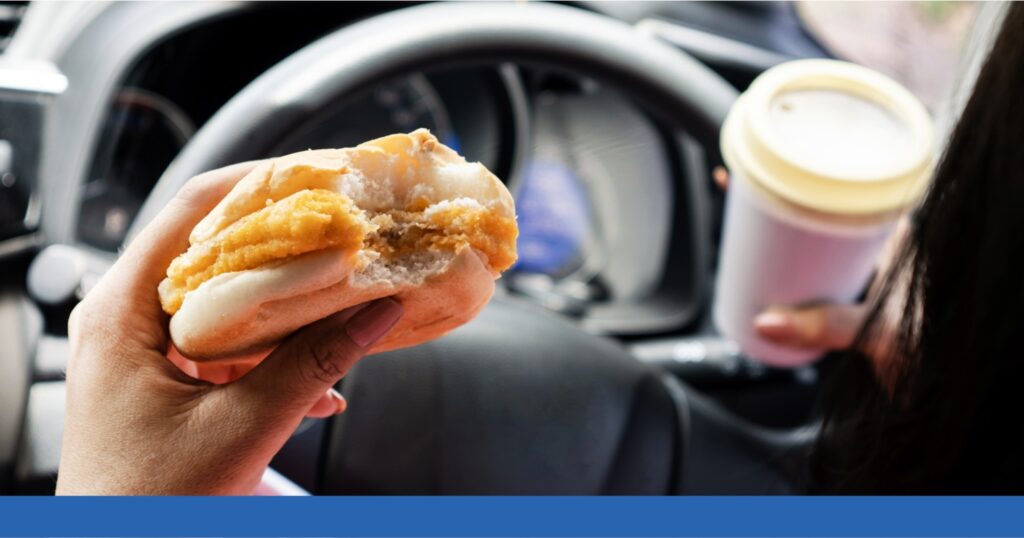 A distracted driver attempts to eat breakfast while behind the wheel of a car.