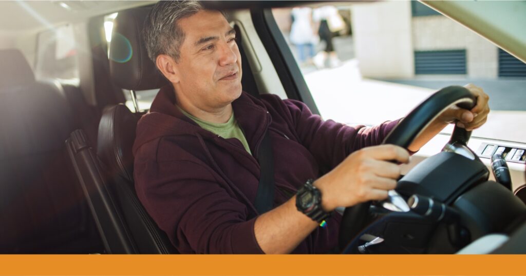 A middle-aged man drives down the road on a sunny day, both hands on the wheel, eyes on the road.