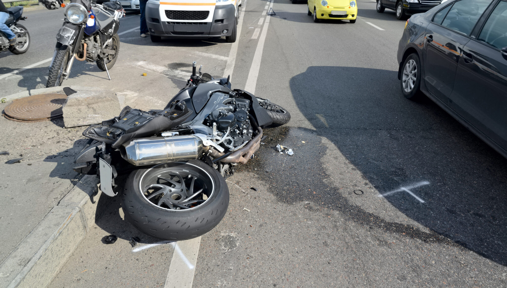 A damaged motorcycle lies on its side at the edge of a city street, with cars stopped nearby and visible debris on the road.