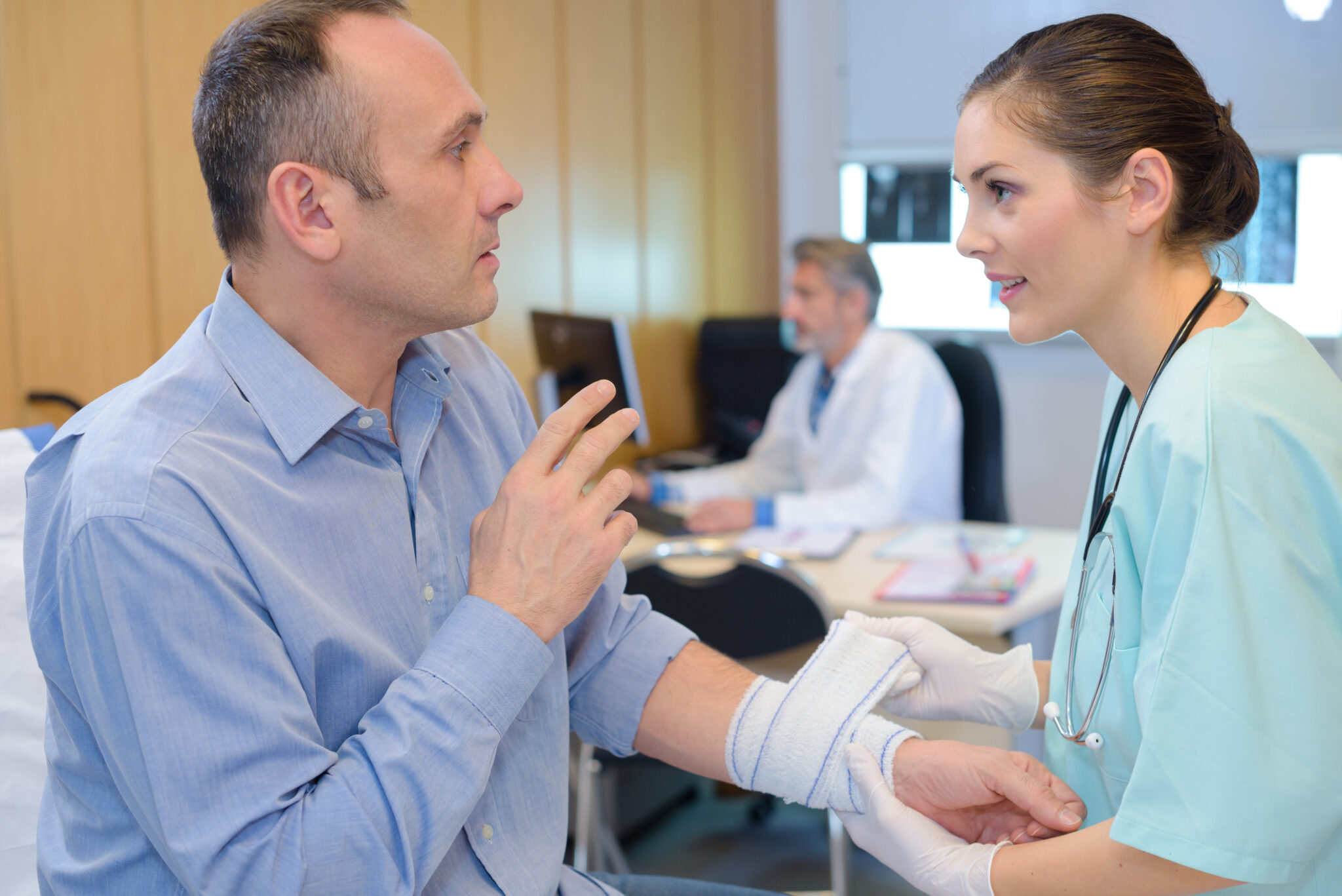 A healthcare professional wearing gloves examines and bandages a man’s injured forearm while they talk inside a clinical setting.