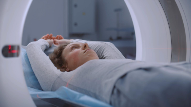 A patient lies on a medical bed with arms resting above their head, entering a CT scanner, with faint laser positioning lines visible