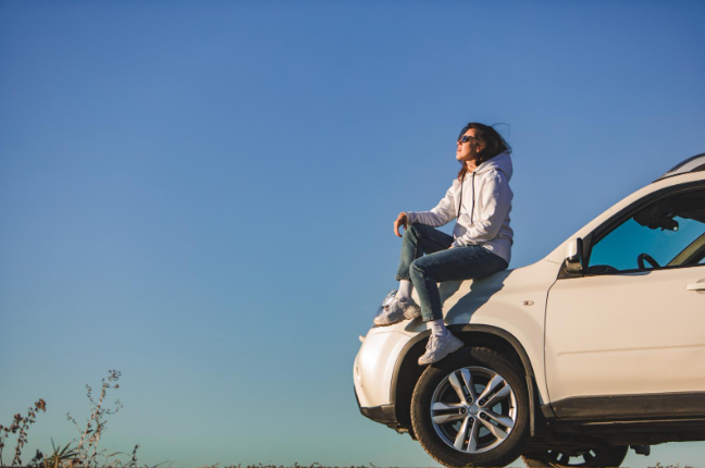 A woman wearing a hoodie and sunglasses sits on the hood of a white SUV, gazing thoughtfully into the distance under a clear blue sky, with a relaxed and contemplative posture.
