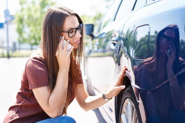 A woman with long hair and glasses crouches next to a car while talking on the phone, inspecting a visible dent or scratch on the side of the vehicle near the wheel.