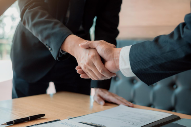 Two business professionals in suits shaking hands over a contract or agreement at a desk, representing partnership or deal-making.