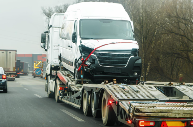 A damaged white delivery van with a crushed front end being transported on a flatbed tow truck along a busy roadway.