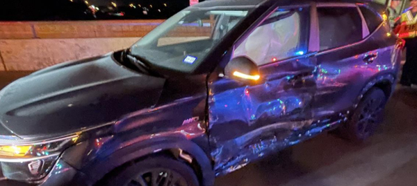 A black SUV with severe damage to the driver's side, including crumpled doors and panels, is parked on a roadway at night with emergency personnel and flashing lights visible in the background.