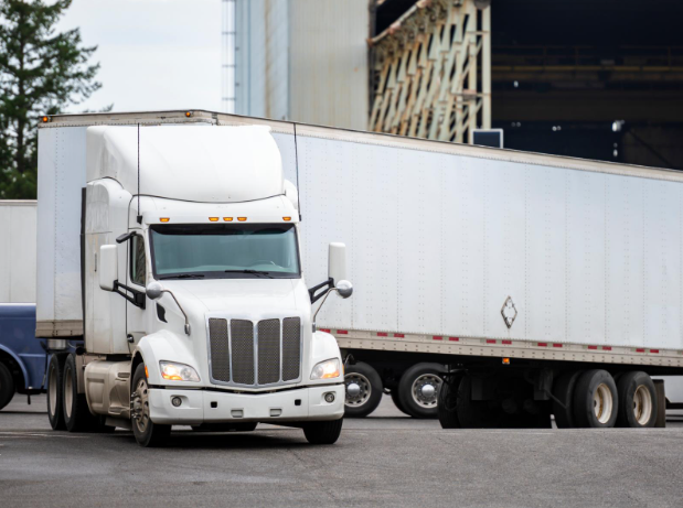 A large white semi-truck with a trailer making a turn in an industrial area, positioned near a warehouse or loading dock.