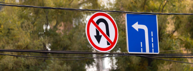 Two traffic signs suspended on wires: one prohibiting U-turns (red circle with a slash over a U-turn arrow), and the other indicating a left-turn-only lane (blue sign with white arrow).