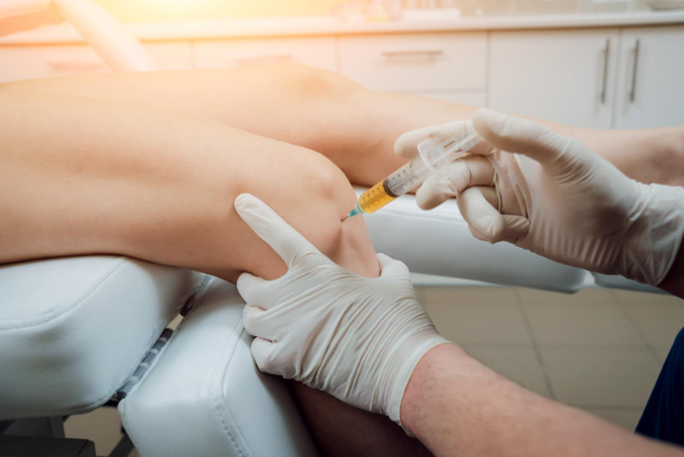 A medical professional wearing gloves injects a syringe into a patient’s knee while the patient lies on an examination chair.