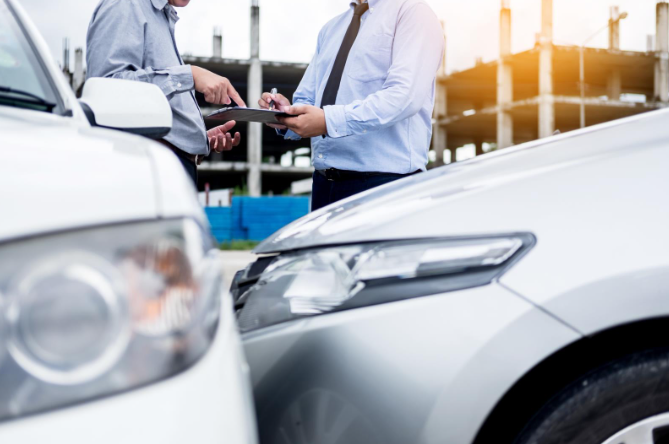 Two men in business attire standing between two cars involved in an accident, reviewing paperwork on a clipboard with a damaged vehicle in the foreground.