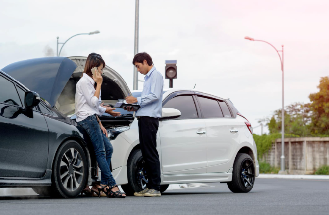A woman on the phone and a man writing on a clipboard stand beside two cars that have collided at an intersection, with one car’s hood open.