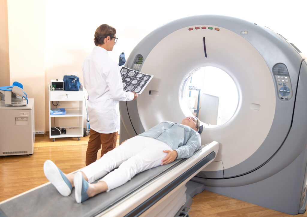 A patient lies on a CT scanner bed while a doctor in a white coat examines brain scan images beside the machine.