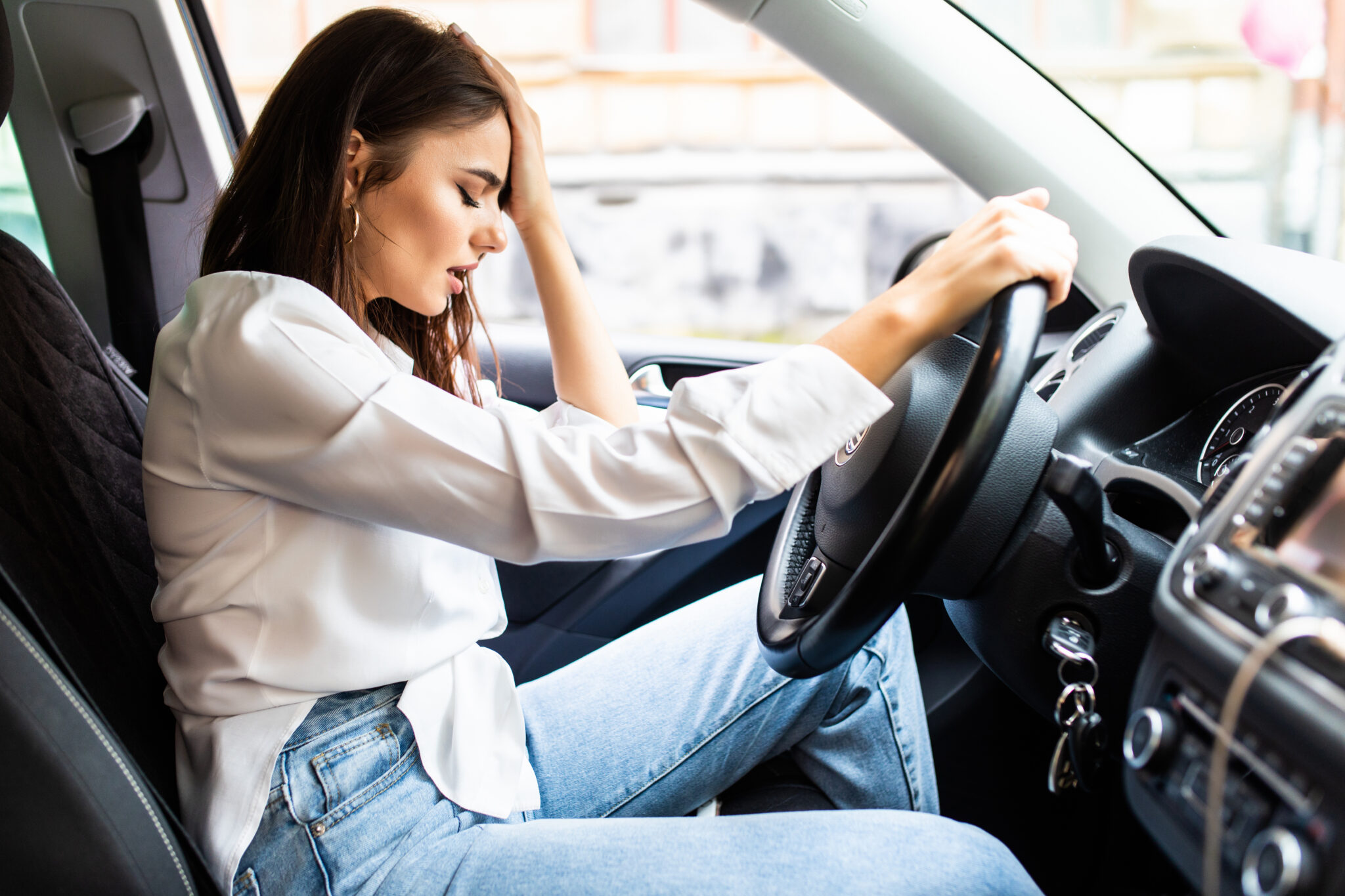 A young woman sits in the driver’s seat of a car, holding her head with one hand and gripping the steering wheel with the other, appearing dizzy or in distress.