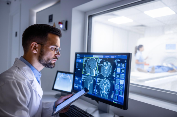 A medical professional in a white coat examines brain scan images on multiple monitors while holding a tablet. In the background, a patient undergoes an MRI scan inside the machine.