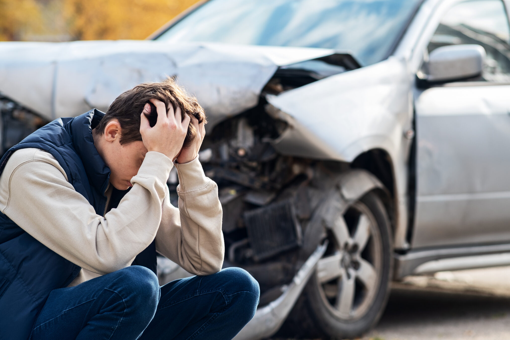 A distressed man crouching near a heavily damaged car after an accident, holding his head in frustration or shock.