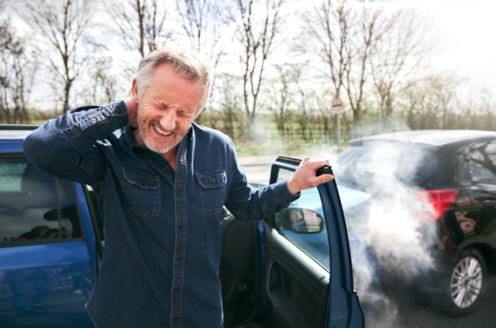 A man standing beside his car after an accident, grimacing and holding his neck in pain, with smoke rising from the vehicle in the background.