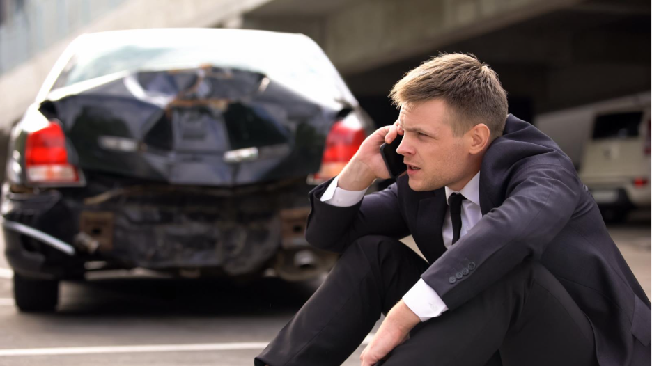 Man sitting near a damaged car while speaking on a phone after a vehicle accident