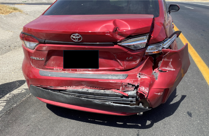 Rear view of a red sedan with significant damage to the trunk and rear bumper, stopped on a roadway after a collision.