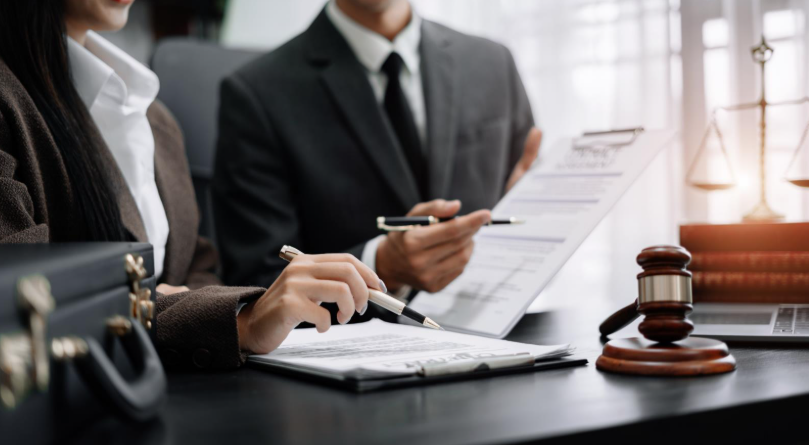Two legal professionals in business attire review and sign documents at a desk, with a judge’s gavel and law books visible nearby.