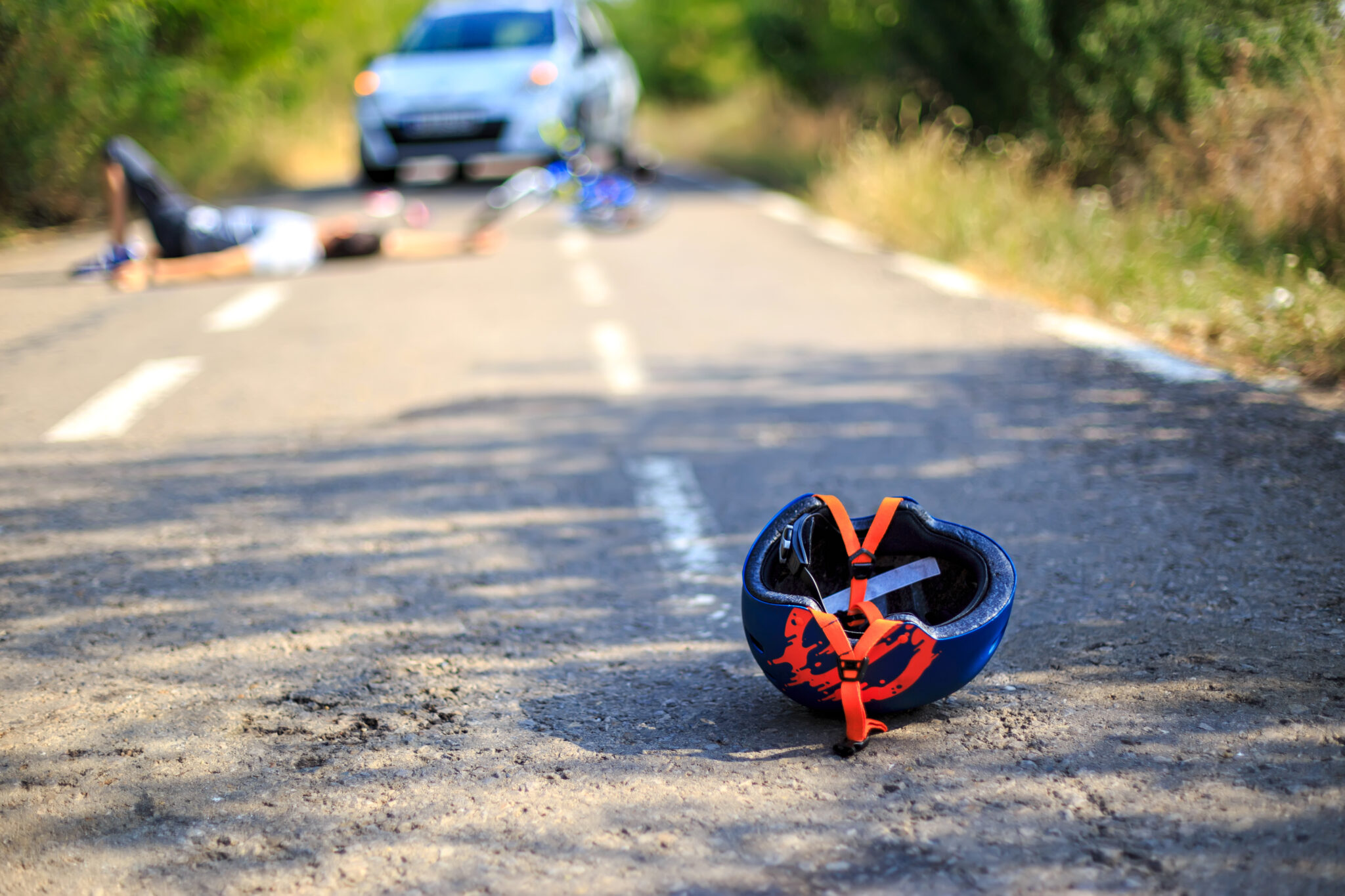 Bicycle helmet lying on the road after an accident, with a cyclist and car blurred in the background.