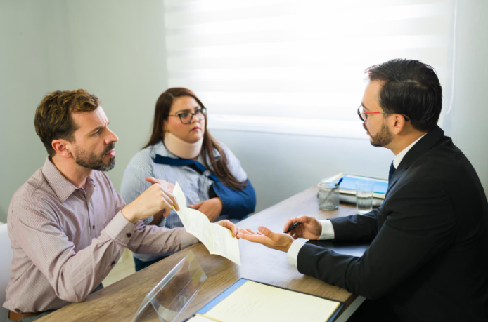 A man in a suit talks with a couple at a desk; the woman wears a neck brace while the man holds paperwork during the meeting.