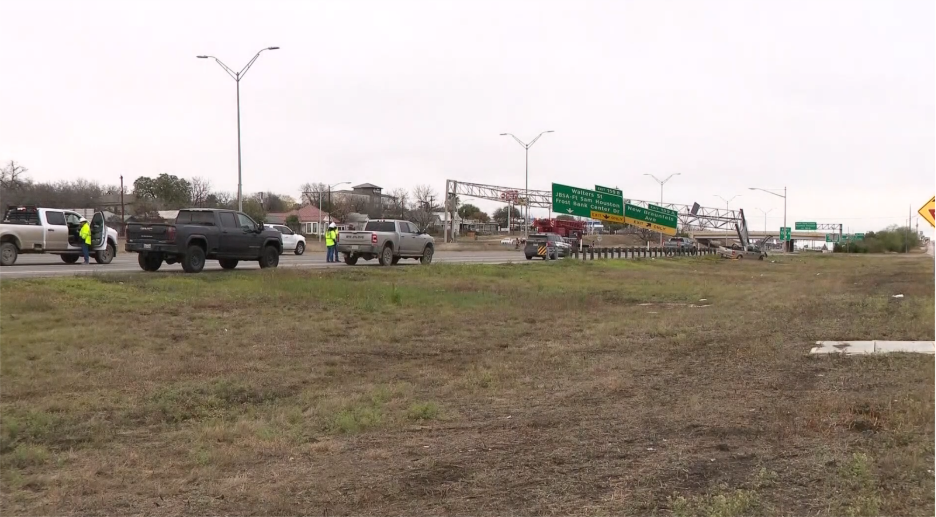 Wide view of a highway accident scene with multiple stopped vehicles, emergency responders, and overhead road signs along a grassy shoulder.
