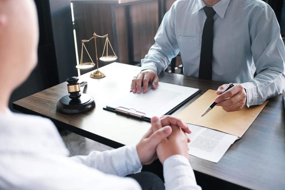 Attorney meeting with a client at a desk, reviewing documents with a pen; a gavel and scales of justice sit on the table.