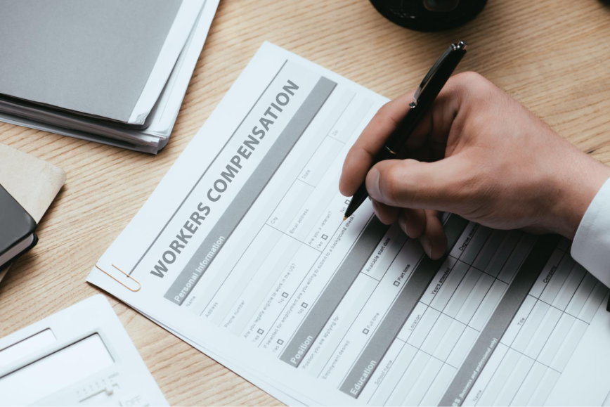 Close-up of a person filling out a “Workers Compensation” claim form on a desk with office supplies nearby.