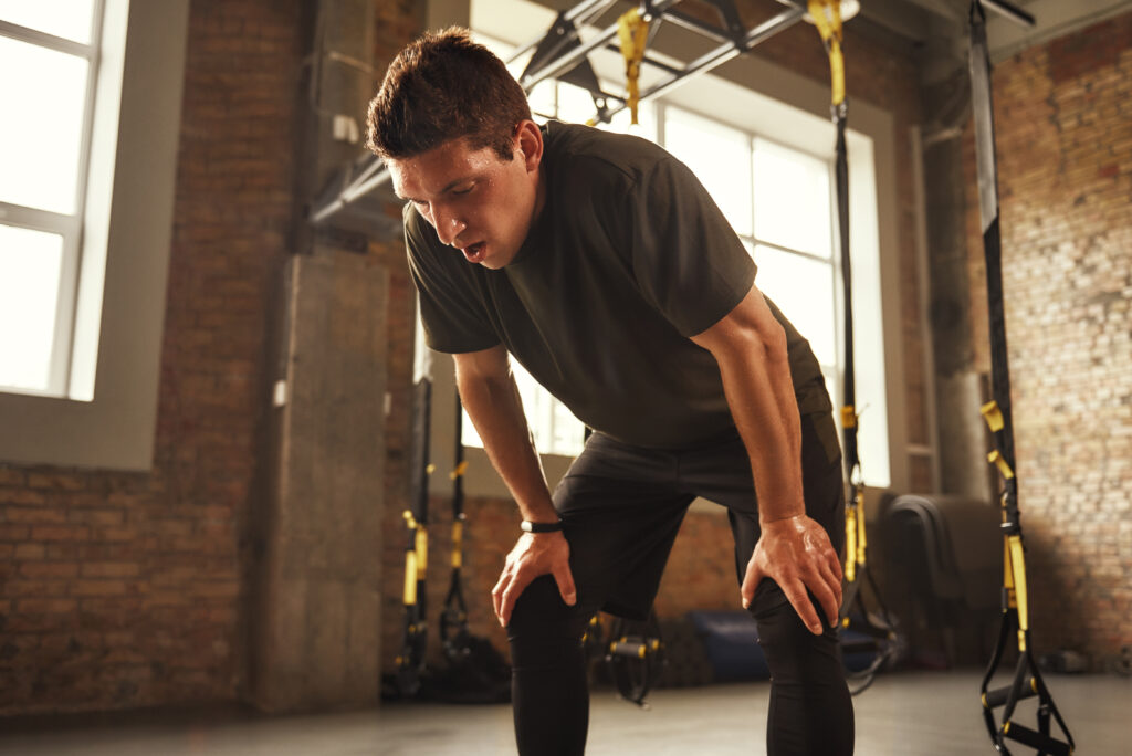 Just a minute to rest. Young athletic man in sportswear looking exhausted while standing at gym. Tiredness. Professional sport. TRX Training.