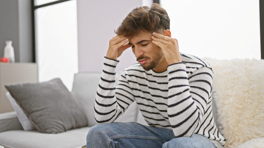 Young man sitting on a couch holding his temples, appearing stressed or in pain, possibly dealing with a headache or emotional distress.