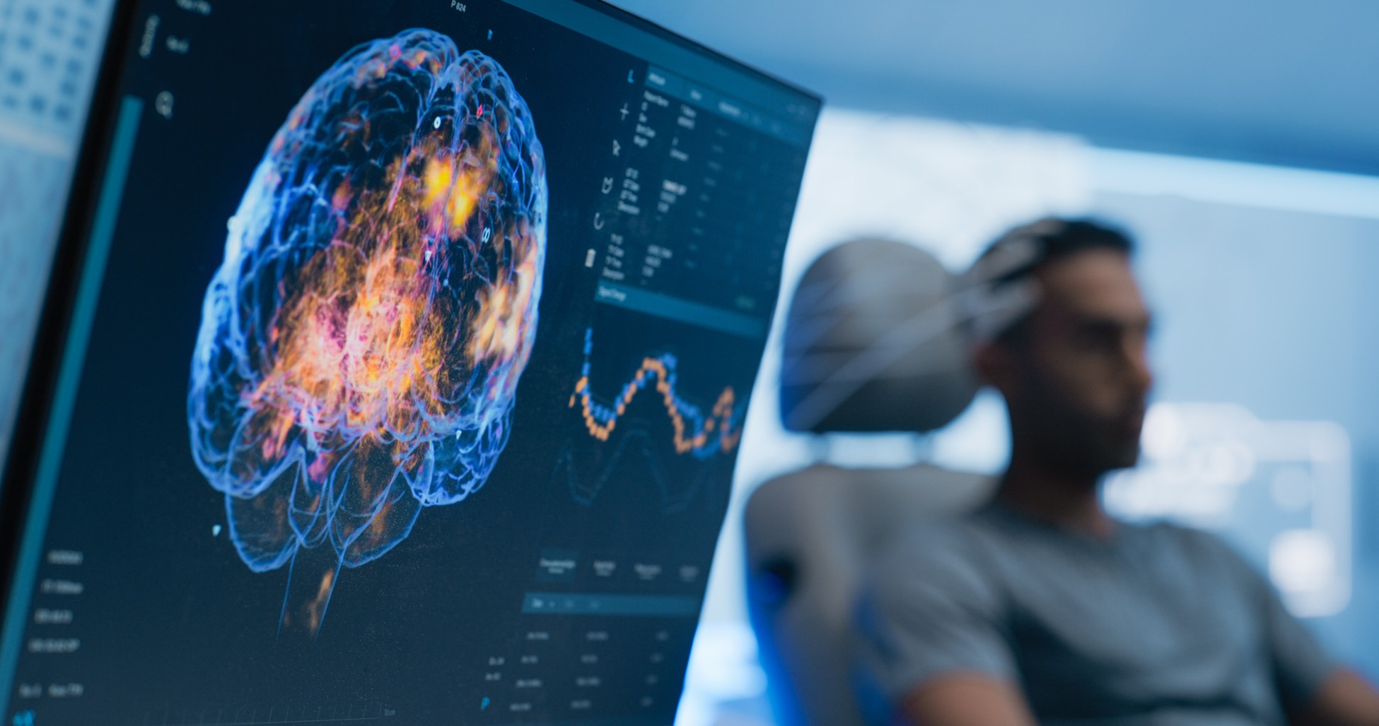 Computer screen displaying a detailed brain scan and data charts while a person undergoes neurological testing in the background.