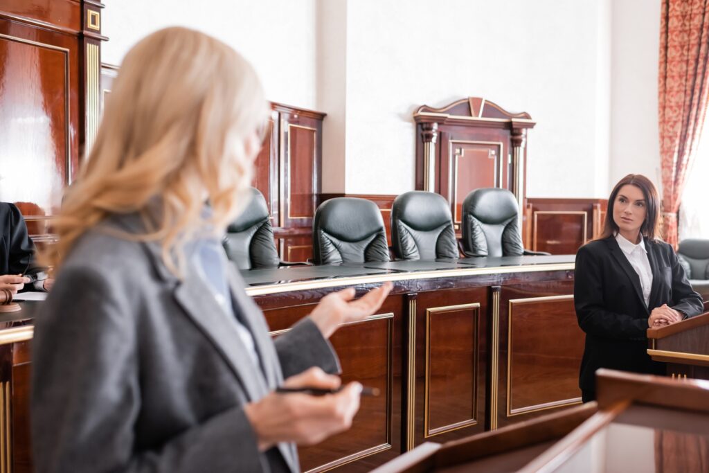 Attorney speaking in a courtroom while another woman stands at the witness stand during a legal proceeding.