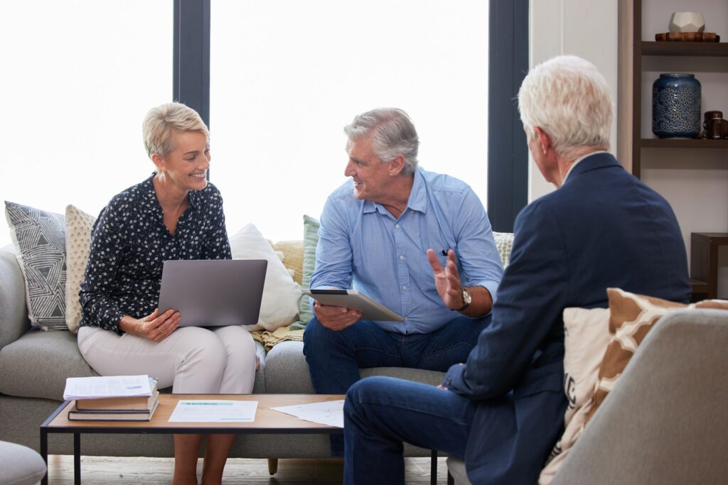 Two adults meeting with a professional advisor in a living room, reviewing information on a laptop and tablet.
