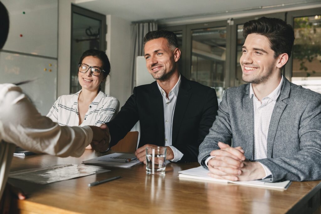 Business professionals smiling and shaking hands across a table during a meeting, signaling a successful agreement or partnership.