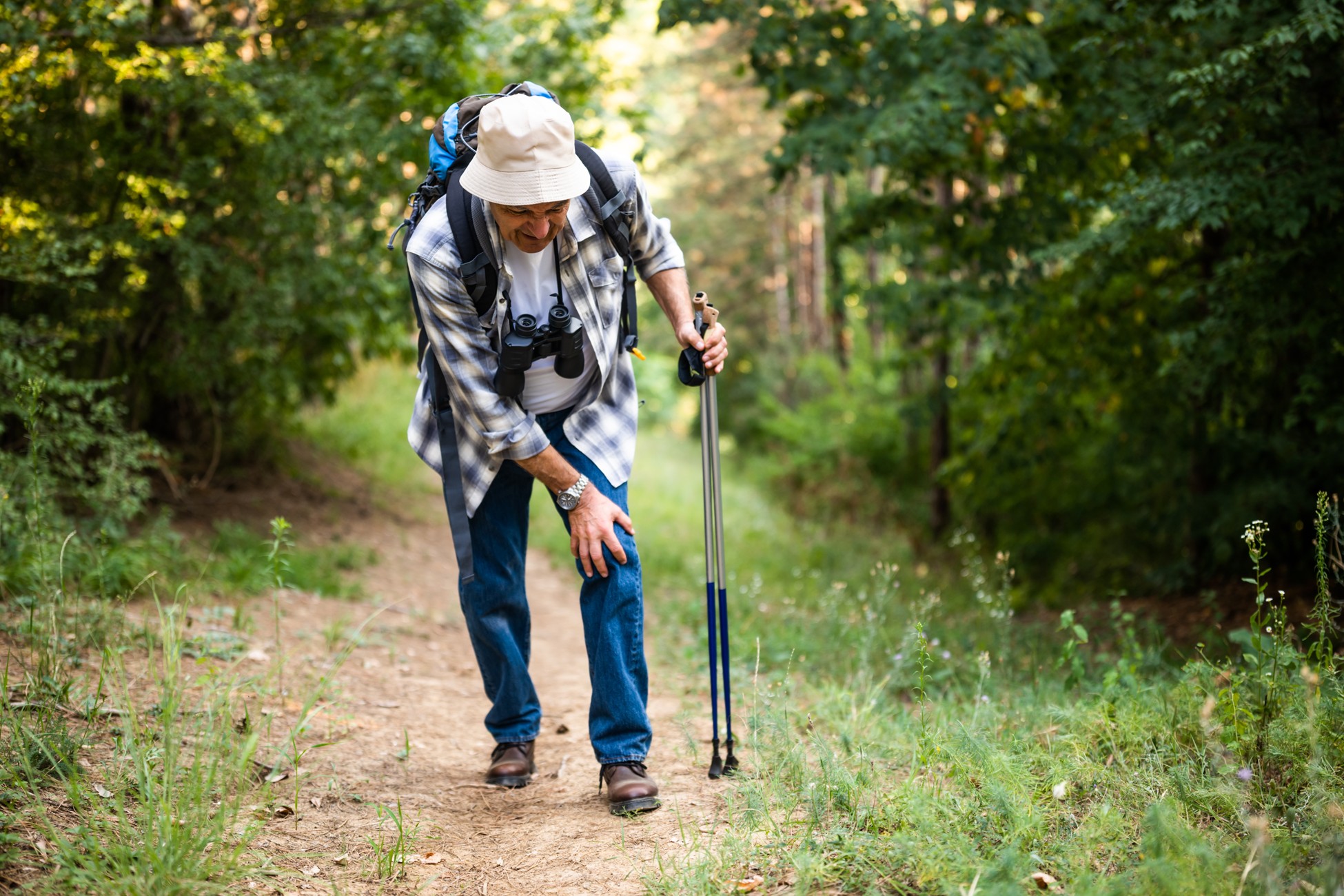 Hiker on a wooded trail holding trekking poles and gripping his knee in discomfort.
