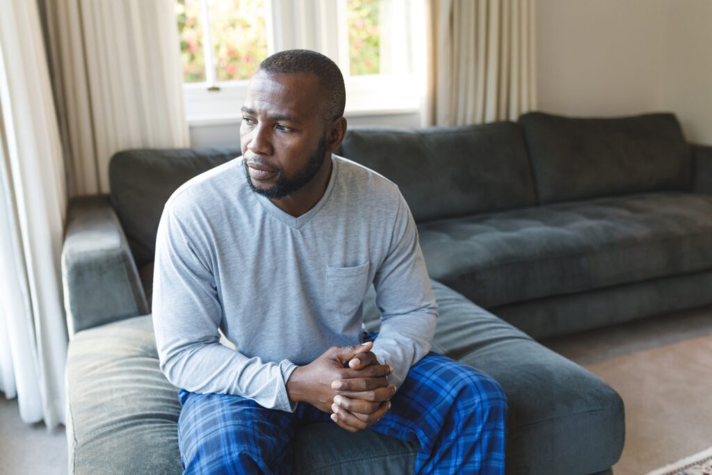 Man sitting on a couch at home with hands clasped, looking off to the side with a thoughtful expression.