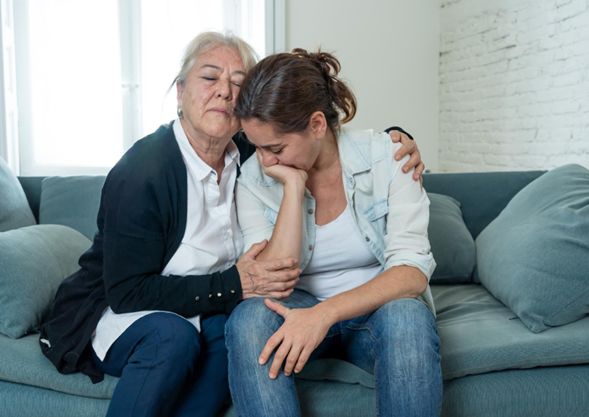 Older man comforting a distressed woman sitting on a gray couch, showing emotional support and compassion in a home setting with bright windows